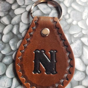 May include: A teardrop-shaped brown leather keychain featuring a stamped letter N in the center. The edges of the leather are decorated with a sunburst-style border pattern. A silver-colored metal rivet secures the top, and a metal ring is attached.