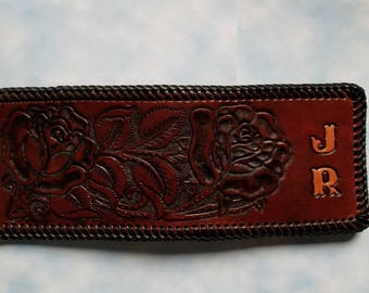 May include: A rectangular brown leather item featuring an embossed floral pattern of two roses with leaves. The edges are finished with a dark braided leather border. The letters J and R are embossed on the right side in a serif font style.