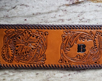 May include: A brown leather wallet with a tooled floral design and a black letter "B" in the center. The wallet has a brown braided border.