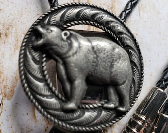 Puede incluir: Un bolo tie plateado con un diseño de oso con un borde de cuerda. El bolo tie está en un cordón trenzado negro.