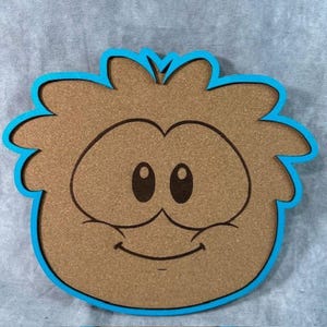 May include: A brown, cartoon-style coaster with a blue border. The coaster features a smiling face with large eyes and a simple mouth. The design is reminiscent of a popular cartoon character.