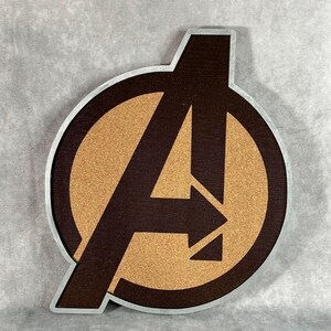 May include: A wooden Avengers logo with a brown background and a silver border. The letter "A" is prominently displayed in a dark brown color, with a tan-colored cork-like texture filling the interior. The logo is circular.