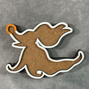 May include: A decorative coaster in the shape of a stylized dog. The coaster has a cork center, a white outline, and a black edge. The dog's nose is accented with an orange ring.
