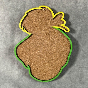May include: A cork coaster with a whimsical design. The coaster has a brown cork center, a green outline, and a yellow accent. The shape is reminiscent of a cartoon character's head. This coaster is designed to protect surfaces from drink condensation.