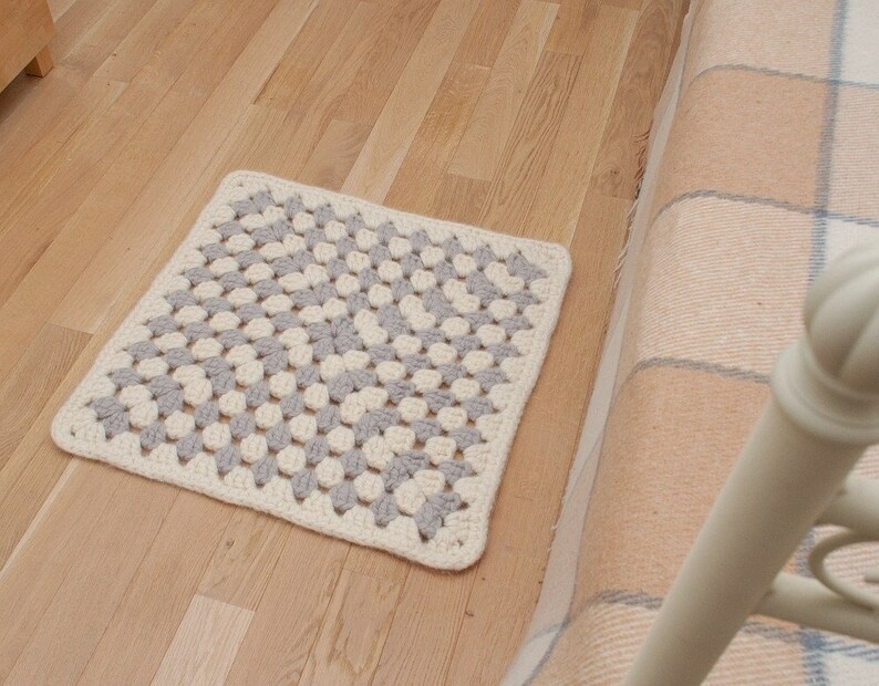 K&ouml;nnte beinhalten: Ein geh&auml;kelter Teppich mit einem quadratischen Muster in Grau- und Wei&szlig;t&ouml;nen. Der Teppich liegt auf einem Holzboden.