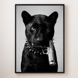 Op de afbeelding: Zwart-wit ingelijste print met een zwarte panter met een halsband met studs, die een zilveren pistool in zijn bek houdt. De intense blik van de panter en het pistool creëren een opvallend contrast.