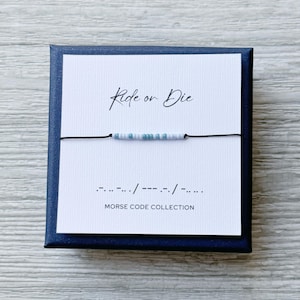 以下が含まれることがあります： 紺色の箱の中に、モールス信号のブレスレットが付いた白いカードが入っています。「Ride or Die」と筆記体で書かれ、モールス信号の翻訳が表示されています。ブレスレットは水色と白のビーズです。「Morse Code Collection」の文字が下にあります。