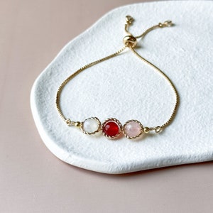 Peut inclure: Un bracelet &agrave; cha&icirc;ne r&eacute;glable dor&eacute; orn&eacute; de trois pierres cabochons rondes dans un sertissage en m&eacute;tal torsad&eacute;. La pierre centrale est rouge fonc&eacute;, flanqu&eacute;e d'une pierre blanche translucide &agrave; gauche et d'une pierre rose p&acirc;le &agrave; droite.