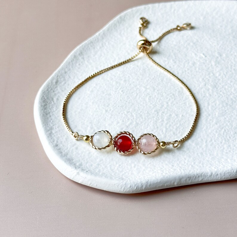 Peut inclure: Un bracelet &agrave; cha&icirc;ne r&eacute;glable dor&eacute; orn&eacute; de trois pierres cabochons rondes dans un sertissage en m&eacute;tal torsad&eacute;. La pierre centrale est rouge fonc&eacute;, flanqu&eacute;e d'une pierre blanche translucide &agrave; gauche et d'une pierre rose p&acirc;le &agrave; droite.