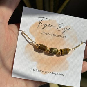 May include: A gold chain bracelet featuring tiger eye crystal beads. The bracelet is displayed on a card with the text "Tiger Eye Crystal Bracelet" and the words "Confidence, Grounding, Clarity."