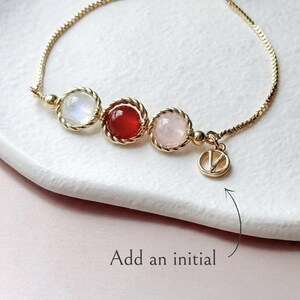 Peut inclure: Un bracelet ajustable dor&eacute; orn&eacute; de trois pierres cabochons rondes blanches, rouges et rose p&acirc;le, chacune sertie dans un cadre en m&eacute;tal torsad&eacute;. Une petite breloque circulaire avec la lettre V pend &agrave; la cha&icirc;ne. Le texte Add an initial est visible.