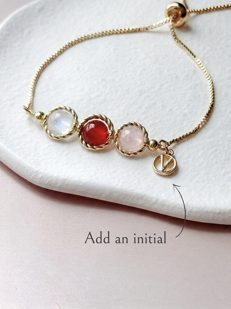 Peut inclure: Un bracelet ajustable dor&eacute; orn&eacute; de trois pierres cabochons rondes blanches, rouges et rose p&acirc;le, chacune sertie dans un cadre en m&eacute;tal torsad&eacute;. Une petite breloque circulaire avec la lettre V pend &agrave; la cha&icirc;ne. Le texte Add an initial est visible.
