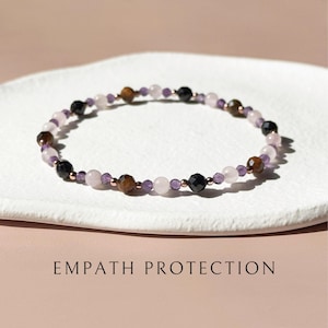 May include: A beaded bracelet featuring alternating round and faceted stones in shades of black, brown, pale pink, and translucent purple, accented with small metallic beads. The text EMPATH PROTECTION is printed below the bracelet.