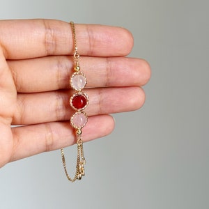 Peut inclure: Un bracelet &agrave; cha&icirc;ne dor&eacute; orn&eacute; de trois breloques circulaires. La breloque centrale contient une pierre rouge fonc&eacute;, tandis que les deux breloques lat&eacute;rales contiennent des pierres rose p&acirc;le. Chaque pierre est sertie dans un cadre en m&eacute;tal dor&eacute; torsad&eacute;.