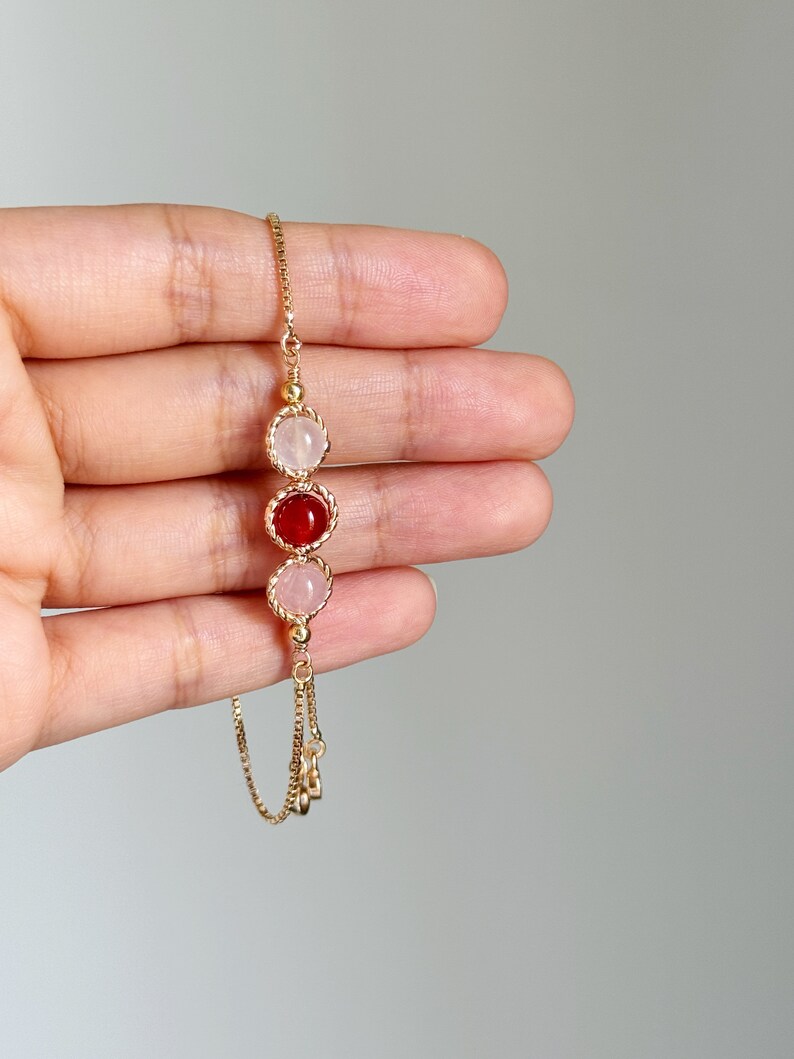 Peut inclure: Un bracelet &agrave; cha&icirc;ne dor&eacute; orn&eacute; de trois breloques circulaires. La breloque centrale contient une pierre rouge fonc&eacute;, tandis que les deux breloques lat&eacute;rales contiennent des pierres rose p&acirc;le. Chaque pierre est sertie dans un cadre en m&eacute;tal dor&eacute; torsad&eacute;.