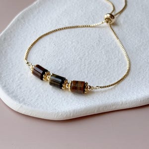 May include: A gold-colored adjustable bracelet featuring three rectangular tiger's eye beads. The beads are a mix of brown and gold tones, with a gold chain and accents. The bracelet is displayed on a white, oval-shaped dish.