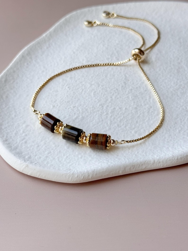 May include: A gold-colored adjustable bracelet featuring three rectangular tiger's eye beads. The beads are a mix of brown and gold tones, with a gold chain and accents. The bracelet is displayed on a white, oval-shaped dish.