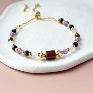 Könnte beinhalten: Ein verstellbares Armband in Goldoptik mit einer zentralen zylindrischen Tigerauge-Perle, flankiert von kleinen blumenförmigen goldenen Zwischenelementen. Das Band besteht aus facettierten Perlen in Violett, Schwarz, Braun und durchscheinendem Weiß, durchsetzt mit kleinen goldenen Rundperlen.