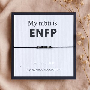 Op de afbeelding: Een witte kaart met zwarte tekst die "My mbti is ENFP" zegt en een zwart koordarmband met zwarte en zilveren kralen. De armband is in morsecode en de tekst onder de kaart zegt "Morse Code Collection".