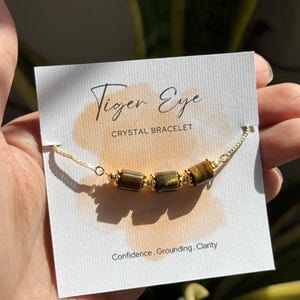May include: A gold chain bracelet with three tiger eye crystal beads. The bracelet is displayed on a white card with the words "Tiger Eye CRYSTAL BRACELET" and the words "Confidence. Grounding. Clarity."