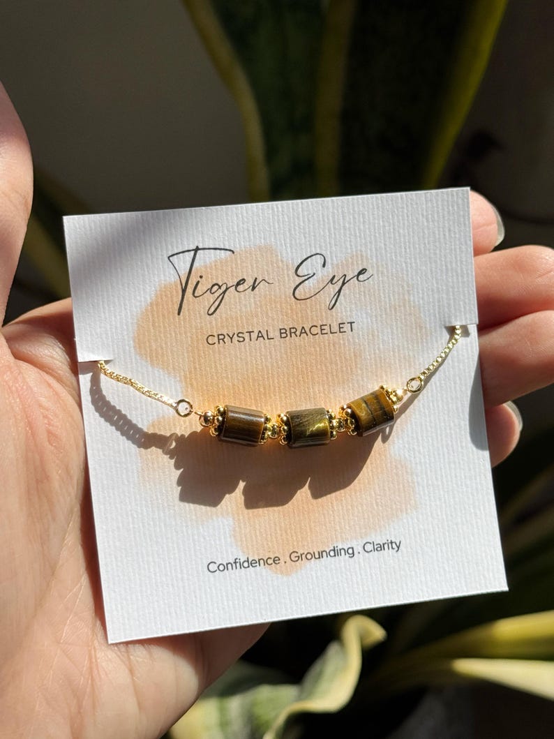 May include: A gold chain bracelet with three tiger eye crystal beads. The bracelet is displayed on a white card with the words "Tiger Eye CRYSTAL BRACELET" and the words "Confidence. Grounding. Clarity."