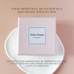 Peut inclure: Une bo&icirc;te &agrave; bijoux carr&eacute;e rose clair avec une &eacute;tiquette blanche centr&eacute;e sur le couvercle. L'&eacute;tiquette pr&eacute;sente une bordure noire et le texte Petite Design ACCESSORY en noir. La bo&icirc;te est pos&eacute;e sur une surface blanche, textur&eacute;e et organique.