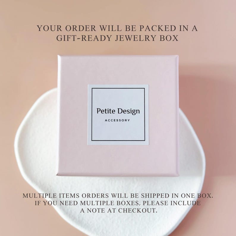 Peut inclure: Une bo&icirc;te &agrave; bijoux carr&eacute;e rose clair avec une &eacute;tiquette blanche centr&eacute;e sur le couvercle. L'&eacute;tiquette pr&eacute;sente une bordure noire et le texte Petite Design ACCESSORY en noir. La bo&icirc;te est pos&eacute;e sur une surface blanche, textur&eacute;e et organique.