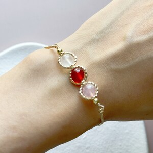 Peut inclure: Un bracelet &agrave; cha&icirc;ne dor&eacute; d&eacute;licat orn&eacute; de trois pierres cabochons rondes serties en ligne. Les pierres comprennent une gemme blanche translucide, une gemme rouge vive et une gemme rose p&acirc;le, chacune entour&eacute;e d'un bord en m&eacute;tal torsad&eacute;.