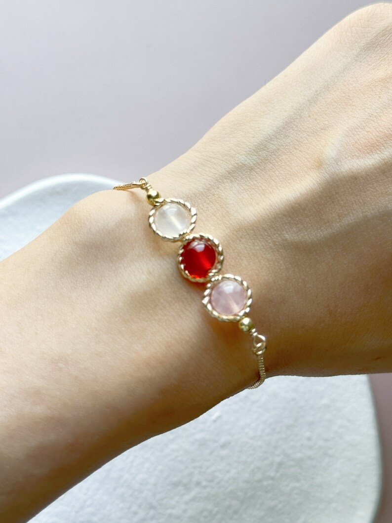Peut inclure: Un bracelet &agrave; cha&icirc;ne dor&eacute; d&eacute;licat orn&eacute; de trois pierres cabochons rondes serties en ligne. Les pierres comprennent une gemme blanche translucide, une gemme rouge vive et une gemme rose p&acirc;le, chacune entour&eacute;e d'un bord en m&eacute;tal torsad&eacute;.