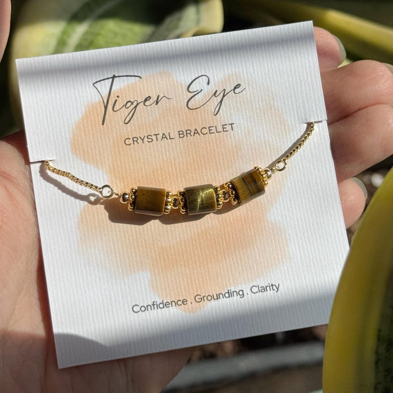 May include: A gold-colored chain bracelet with three rectangular tiger eye stones. The bracelet is displayed on a white card with the words "Tiger Eye CRYSTAL BRACELET" and the words "Confidence, Grounding, Clarity".