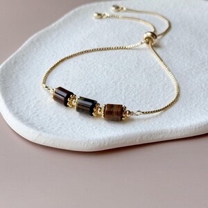 May include: A gold-toned adjustable bracelet featuring three rectangular, brown tiger eye beads. The bracelet has a delicate chain and a sliding bead closure. The beads are accented with small gold-colored beads.