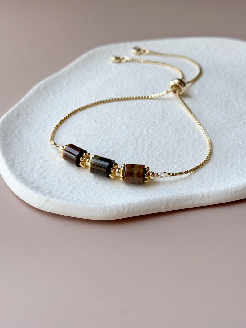 May include: A gold-toned adjustable bracelet featuring three rectangular, brown tiger eye beads. The bracelet has a delicate chain and a sliding bead closure. The beads are accented with small gold-colored beads.