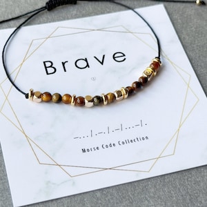 Könnte beinhalten: Ein verstellbares schwarzes Armband mit Tigeraugenperlen und goldfarbenen Akzenten. Das Armband wird auf einer Karte mit dem Wort "Brave" und Morsecode präsentiert. Die Karte trägt auch die Aufschrift "Morse Code Collection".