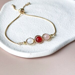 Peut inclure: Un bracelet r&eacute;glable dor&eacute; dot&eacute; d'une cha&icirc;ne fine et de trois pierres rondes serties en ligne. Les pierres se composent d'une perle blanche translucide, d'une perle rouge vif et d'une perle rose p&acirc;le, chacune encadr&eacute;e par un bord en m&eacute;tal torsad&eacute;.