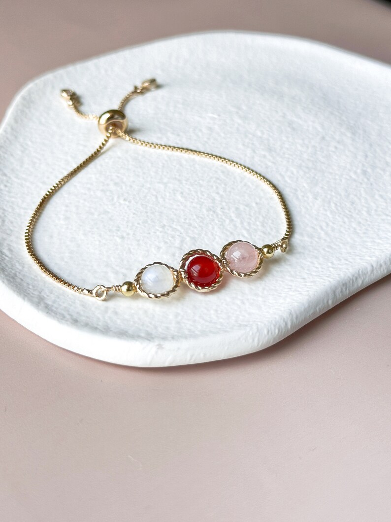 Peut inclure: Un bracelet r&eacute;glable dor&eacute; dot&eacute; d'une cha&icirc;ne fine et de trois pierres rondes serties en ligne. Les pierres se composent d'une perle blanche translucide, d'une perle rouge vif et d'une perle rose p&acirc;le, chacune encadr&eacute;e par un bord en m&eacute;tal torsad&eacute;.