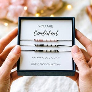 May include: A dark blue gift box containing three thin black cord bracelets with small beads arranged in Morse code patterns. The white card inside features the text YOU ARE Confident and MORSE CODE COLLECTION with a Morse code sequence.