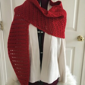 May include: A red crocheted scarf with a lacy pattern. The scarf is draped over a white sweater on a mannequin.