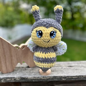 May include: A handmade crochet bee doll with a yellow and gray body, large blue eyes, and small antennae. The bee has white and green wings and is displayed on a wooden stand. The background is blurred green foliage.
