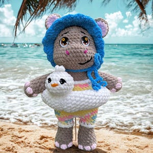 May include: A crocheted hippo doll wearing a blue hat and swim trunks, holding a white duck-shaped floatie. The doll is on a sandy beach with the ocean in the background. The hippo has pink accents on its ears, feet, and cheeks.