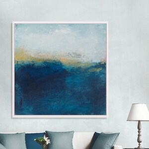 May include: A framed abstract painting with a blue and gold color scheme. The painting depicts a landscape with a horizon line and a sky. The painting is hanging on a wall above a white couch.