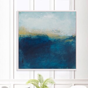 May include: Abstract painting with a blue and gold color scheme. The painting depicts a landscape with a horizon line and a sky with clouds. The painting is framed in a white frame and is hanging on a white wall.