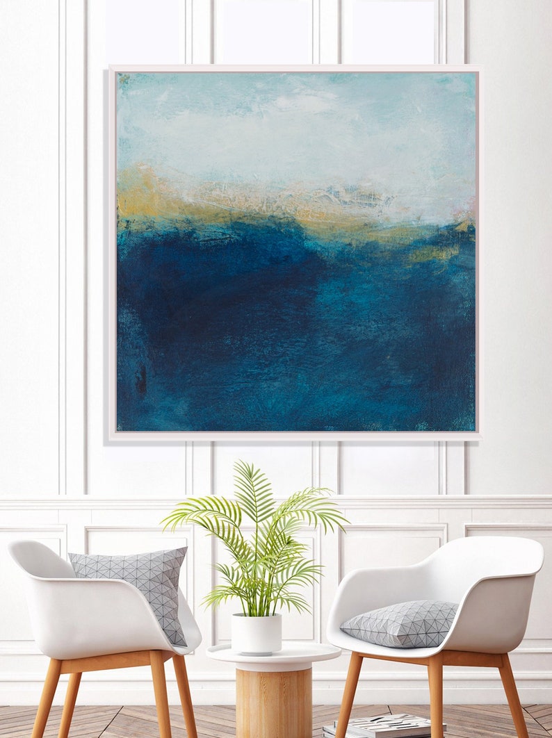 May include: Abstract painting with a blue and gold color scheme. The painting depicts a landscape with a horizon line and a sky with clouds. The painting is framed in a white frame and is hanging on a white wall.