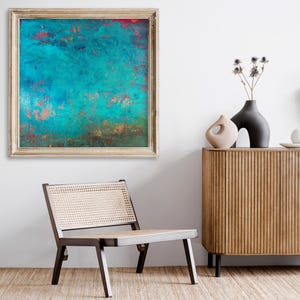 May include: Abstract teal and copper-toned artwork in a gold frame hangs on a white wall. A modern wooden cabinet with black legs and decorative vases sits nearby. A mid-century modern chair with a woven seat is in the foreground.