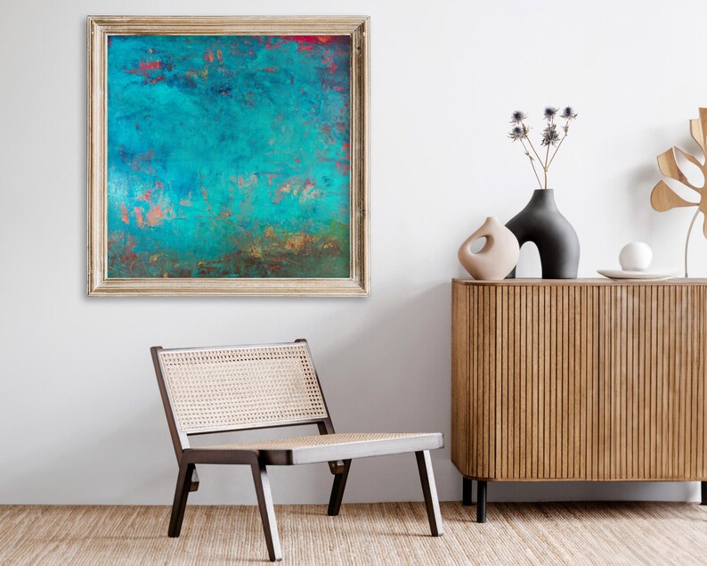 May include: Abstract teal and copper-toned artwork in a gold frame hangs on a white wall. A modern wooden cabinet with black legs and decorative vases sits nearby. A mid-century modern chair with a woven seat is in the foreground.