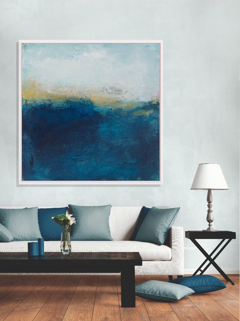 May include: A framed abstract painting with a blue and gold color scheme. The painting depicts a landscape with a horizon line and a sky. The painting is hanging on a wall above a white couch.