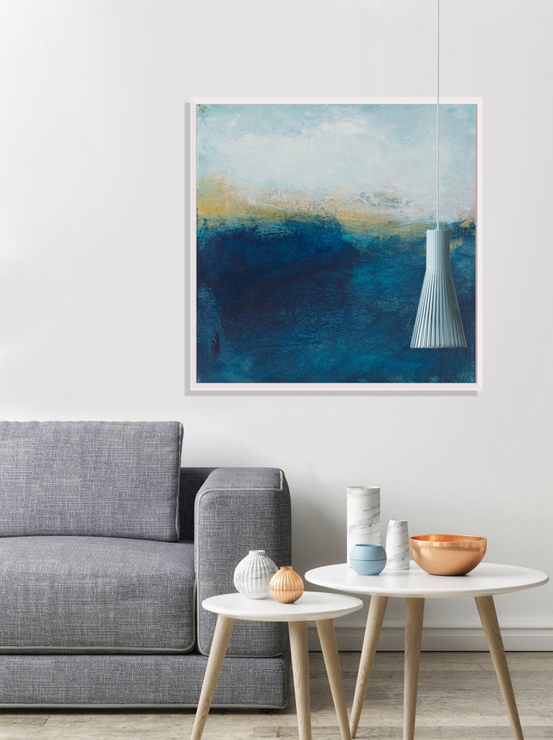 May include: A framed abstract painting with a blue and white color scheme. The painting is hanging on a white wall above a gray sofa and two white coffee tables with decorative objects.