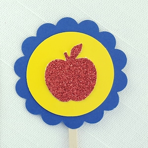 May include: A red glitter apple silhouette on a yellow circle with a blue scalloped border. The apple is on a wooden stick.