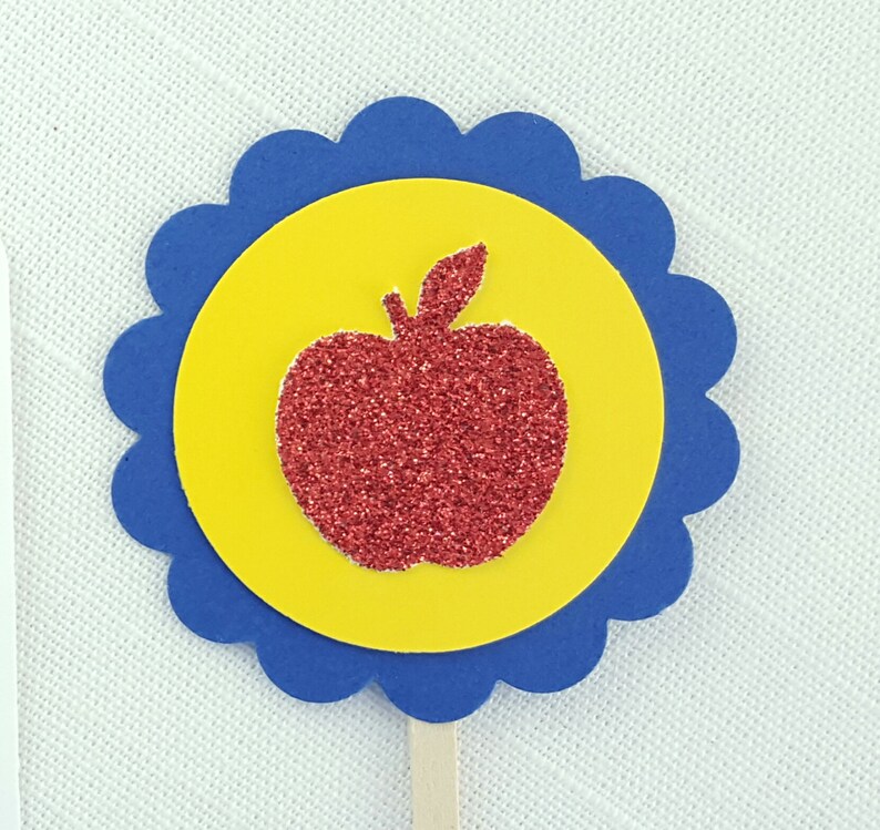 May include: A red glitter apple silhouette on a yellow circle with a blue scalloped border. The apple is on a wooden stick.