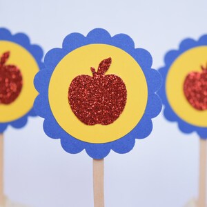 May include: Three red glitter apple cupcake toppers with yellow and blue scalloped circles. The toppers are on wooden sticks.