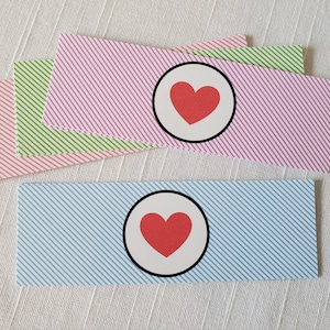 May include: Two gift tags with diagonal stripes. One tag is pink with a black circle and a red heart inside. The other tag is blue with a black circle and a red heart inside.
